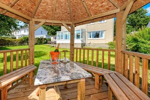 A wooden deck with a table and benches, a pergola overhead, and a view of houses in the background. - 3 Bed in Llanbedrog (oc-glensi) (Llanbedrog)