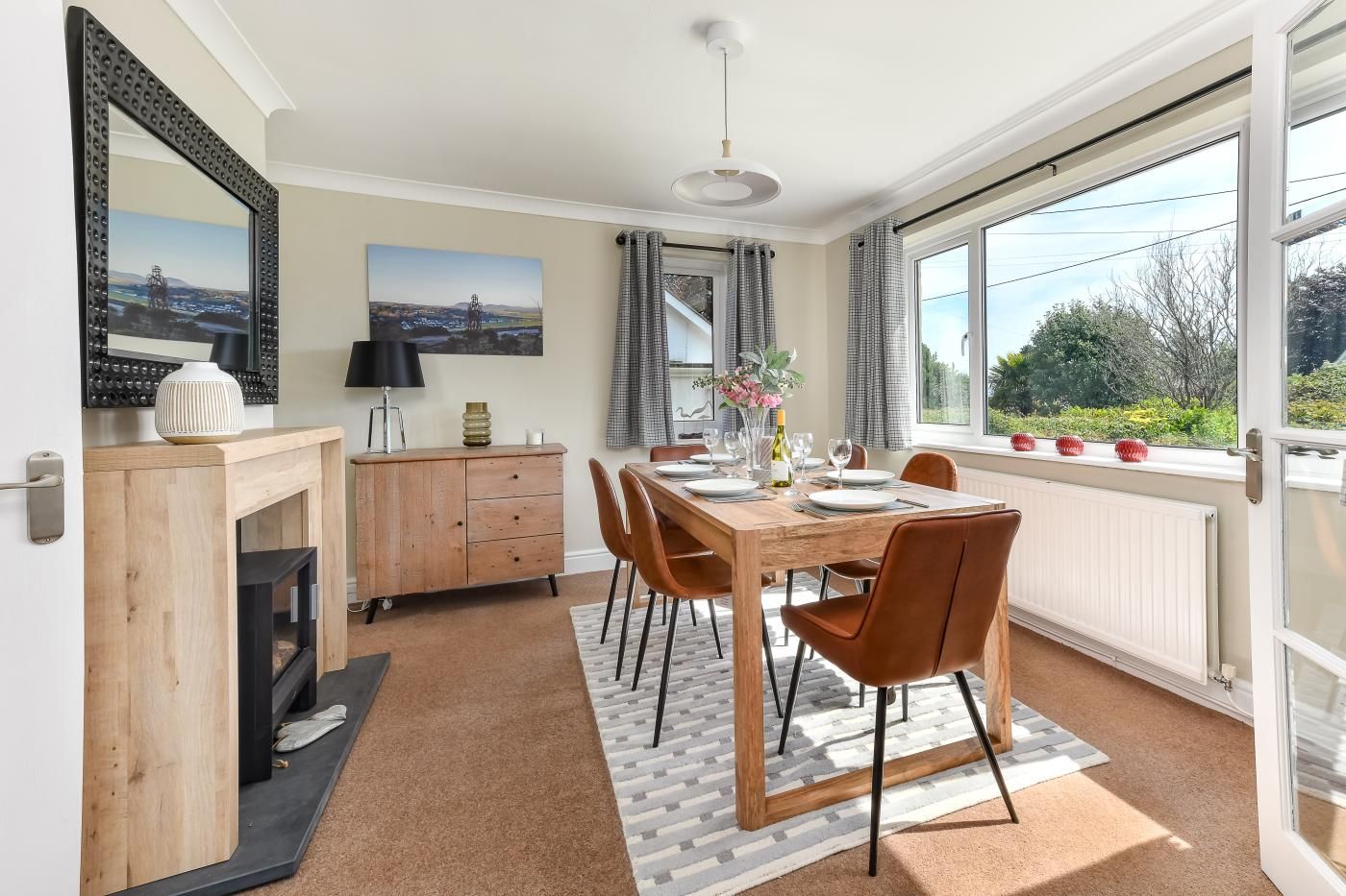 A dining area with a wooden table set for four, a cabinet with drawers, a fireplace, and a large window with curtains.