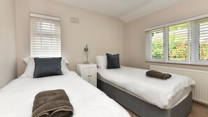 Ein Schlafzimmer mit zwei Betten, jeweils mit weißer Bettwäsche und dunklen Kissen, einem Nachttisch, einer Lampe und einem Fenster mit Jalousien.