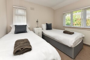 A bedroom with two beds, each with white linens and dark pillows, a nightstand, a lamp, and a window with blinds. - 3 Bed in Llanbedrog (oc-glensi) (Llanbedrog)