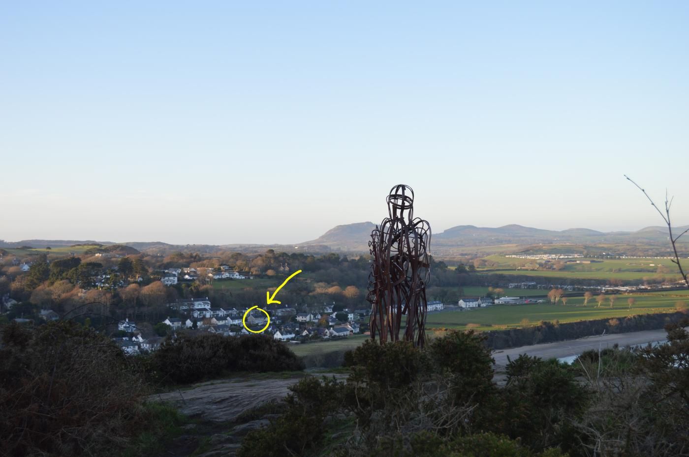 Un paysage rural avec une sculpture métallique au premier plan, un village au milieu et des collines ondulantes à l’arrière-plan.
