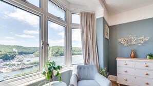 Quarto com vista para um rio e colinas, um aparador branco, uma poltrona cinzenta e uma pequena mesa com uma planta.