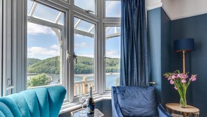 Quarto com uma grande janela que oferece uma vista panorâmica de um rio e colinas, uma poltrona azul, uma mesa pequena com uma garrafa e um copo, e um vaso com flores.