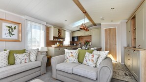 A cozy living room with two sofas and a dining table, a kitchen with wooden cabinets, and a large window with blinds. - 2 Bed in Abersoch (oc-atacam) (Abersoch)
