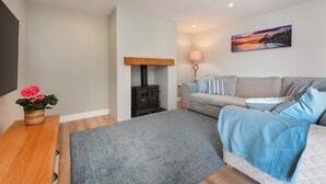 Ein gemütliches Wohnzimmer mit einem grauen Sofa, einem Holztisch mit einer Pflanze, einem schwarzen Kaminofen und einem Bild an der Wand.
