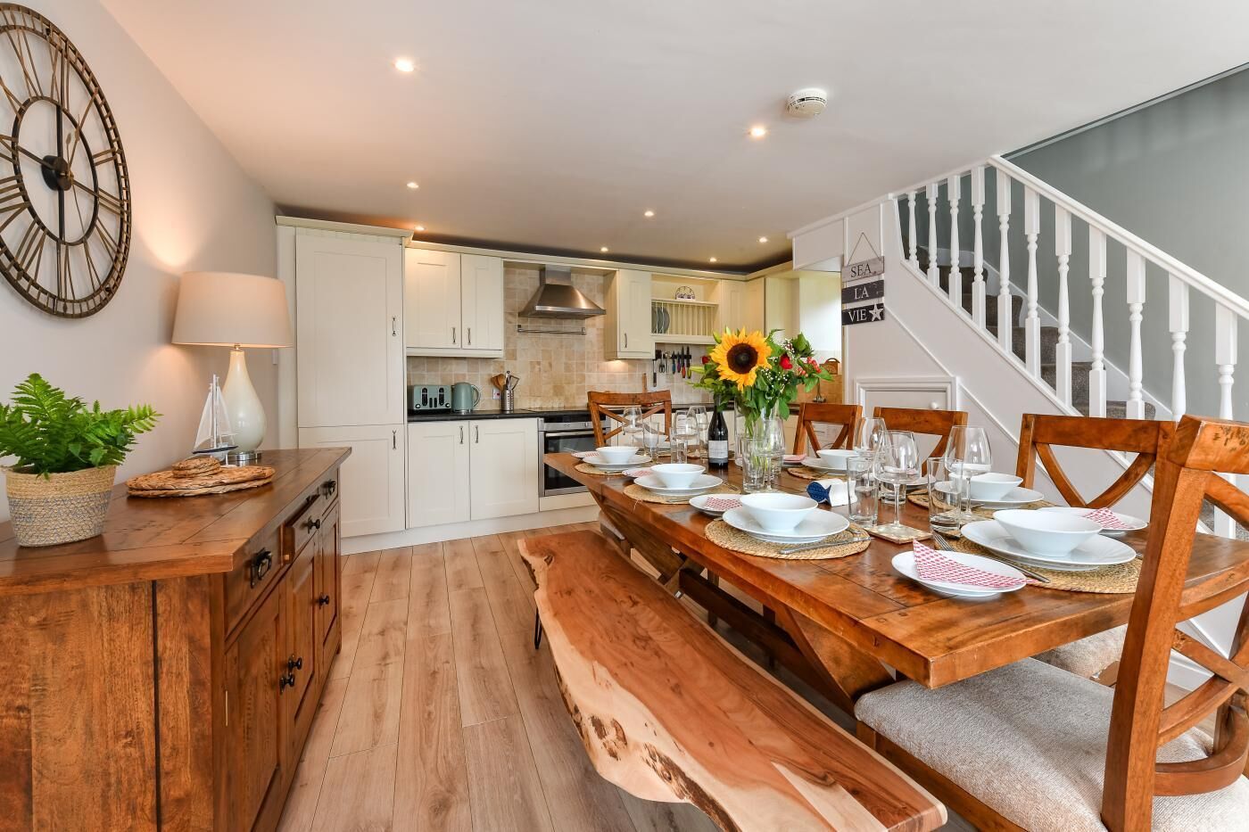 A dining area with a wooden table set for six, white cabinets, a large wall clock, and a staircase leading to another level.
