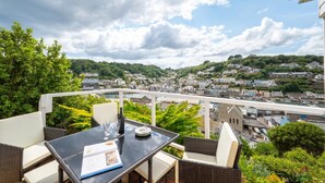A balcony with a table set for two, overlooking a coastal town with a bridge and greenery. - 2 Bed in Looe (oc-mizzen) (Looe)