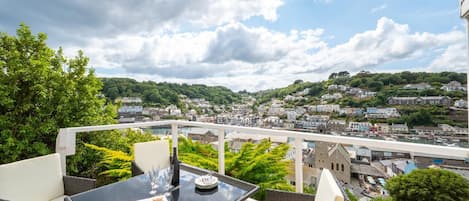 A balcony with a table set for two, overlooking a coastal town with a bridge and greenery.