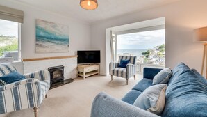 A living room with a fireplace, a television, a sofa, and a chair. There is a painting on the wall and a view of the sea through the window. - 2 Bed in Looe (oc-msweet) (Looe)