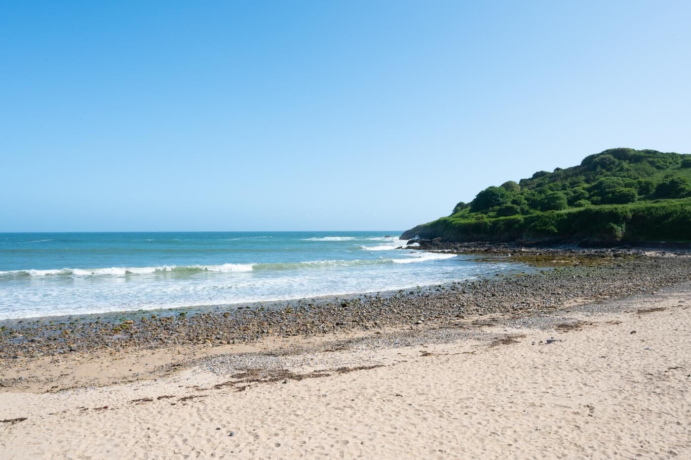 A beach with pebbles and sand, a rocky outcrop with greenery, and the ocean.