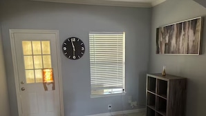 Ein Zimmer mit einer Tür, einer Wanduhr, einem Fenster mit Jalousien, einem Holzkonsolentisch und einem Gemälde an der Wand.