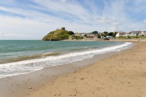 Ein Sandstrand mit sanften Wellen, eine historische Burg auf einem Hügel und eine Reihe von Häusern im Hintergrund.