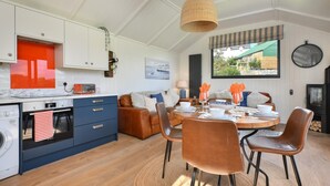 A modern kitchen with white cabinets, a built-in oven, and a dining area with a round table and brown chairs. - 2 Bed in Abererch (oc-glwysf) (Abererch)