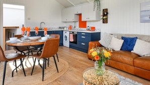 A modern kitchen with white cabinets, a dining area with a round table and chairs, a living room with a sofa and cushions, and a small side table with a vase of flowers. - 2 Bed in Abererch (oc-glwysf) (Abererch)
