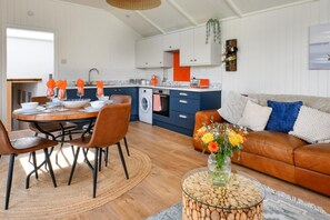 Une cuisine moderne avec des meubles blancs, un coin repas avec une table ronde et des chaises, un salon avec un canapé et des coussins, et une petite table d’appoint sur laquelle est posé un vase de fleurs.