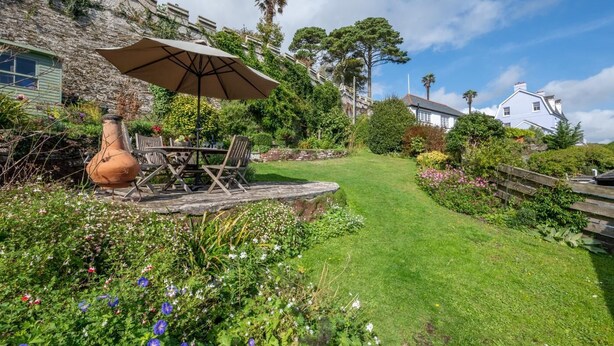 Un jardín con mesa y sillas bajo una sombrilla, rodeado de exuberante follaje y flores.
