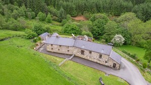 A stone building with a sloped roof, surrounded by lush green fields and dense forest. - 6 Bed in Machynlleth (oc-bow374) (Glantwymyn)