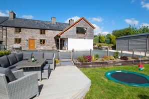 Ein Steinhhaus mit einem Holzanbau, eine Terrasse mit Gartenmöbeln und ein Trampolin.