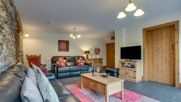 A living room with a black leather sofa, a wooden coffee table, a red patterned rug, a television, and a stone wall. - 6 Bed in Machynlleth (oc-bow374) (Glantwymyn)