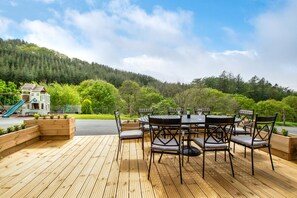 A wooden deck with a round table and chairs, surrounded by a forested hillside. - 6 Bed in Machynlleth (oc-bow374) (Glantwymyn)