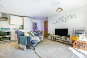 A modern living room with a sofa, a flat-screen TV, a fireplace, and a kitchen area with an oven. - 1 Bed in Fowey (oc-saila) (Fowey)