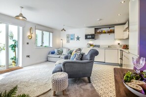 A modern living room with a grey sofa, a small ottoman, and a dining table with a glass of wine. - 1 Bed in Fowey (oc-saila) (Fowey)