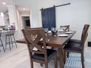 A dining area with a wooden table and chairs, a kitchen in the background, and a blue curtain on the wall. - Westport Reserve Vacation Home w/ Private Pool | Family Friendly (Naples)