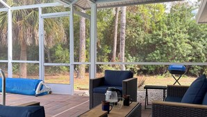 Ein überdachter Patio-Bereich mit Rattanmöbeln, einem Grill und Blick auf die umliegende Bepflanzung.
