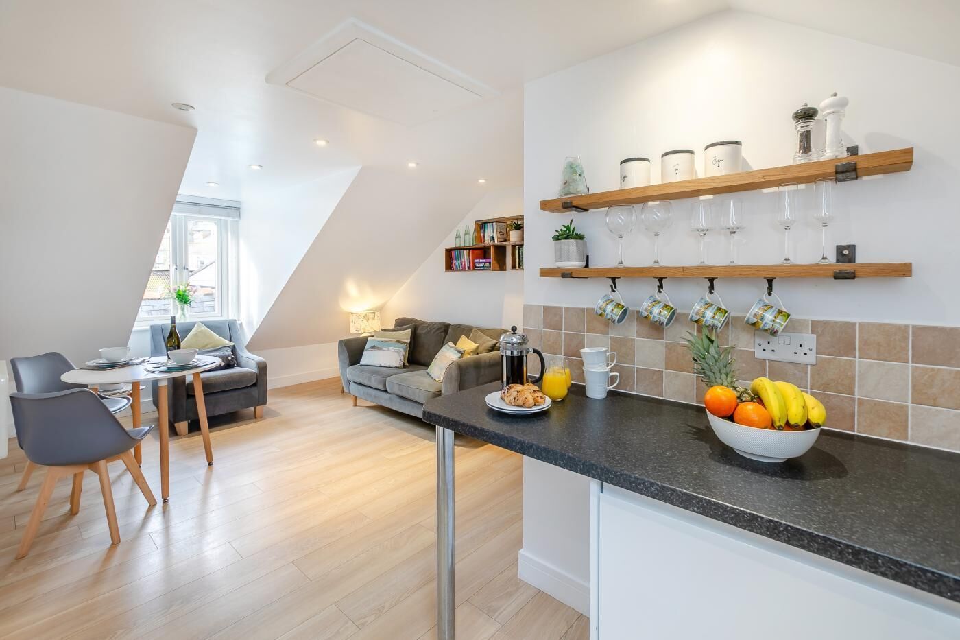 A modern kitchen with a breakfast bar, a dining area with a sofa and chairs, and a kitchen island with fruit and a coffee maker.