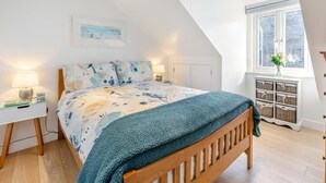 A bedroom with a sloped ceiling, a bed with a patterned bedspread, bedside tables with lamps, a window with a view, and a small cabinet with baskets. - 1 Bed in St. Ives (oc-h27240) (St Ives)