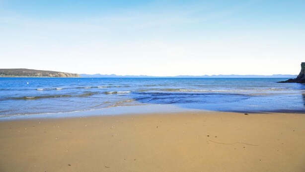 Une plage à l’eau claire, avec un rivage sablonneux et des collines qui se dessinent à l’horizon.