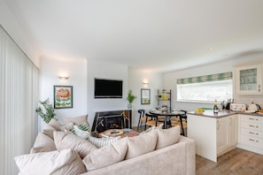 Una sala de estar moderna con sofá, chimenea, comedor con mesa y sillas, y cocina con armarios y ventana.