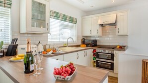Une cuisine moderne avec des armoires blanches, un four encastré, un four à micro-ondes et un plan de travail sur lequel se trouvent une bouteille de vin et un saladier rempli de fruits.