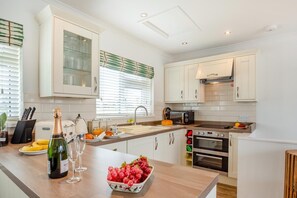 A modern kitchen with white cabinets, a built-in oven, a microwave, and a countertop with a bottle of wine and a bowl of fruit. - 2 Bed in Abersoch (oc-s27221) (Abersoch)