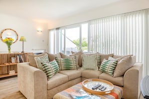 A living room with a sectional sofa, a coffee table with remote controls, a bookshelf, and a window with blinds. - 2 Bed in Abersoch (oc-s27221) (Abersoch)