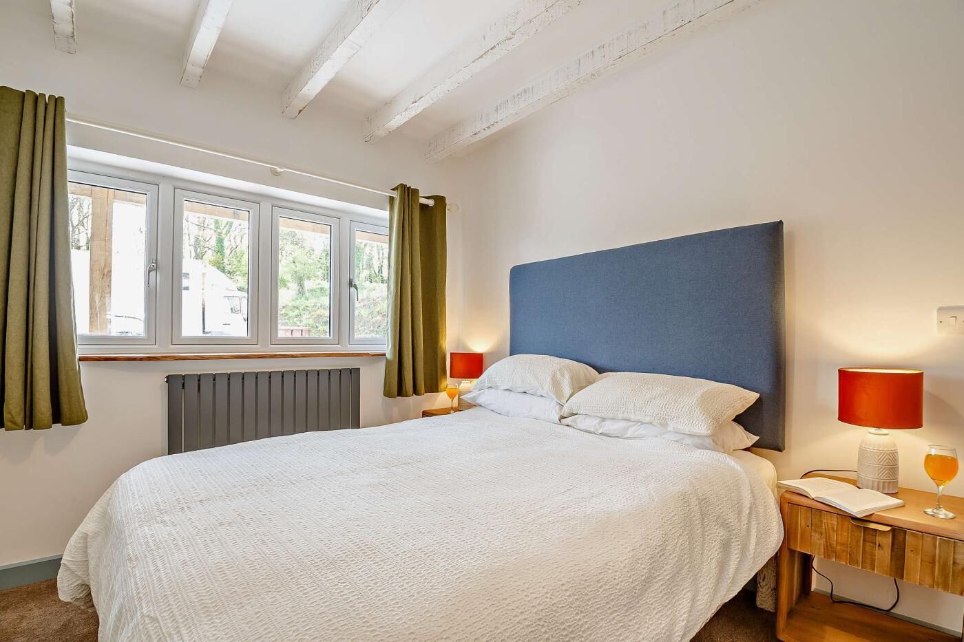 A bedroom with a large bed, two bedside tables with lamps, a window with curtains, and a radiator.