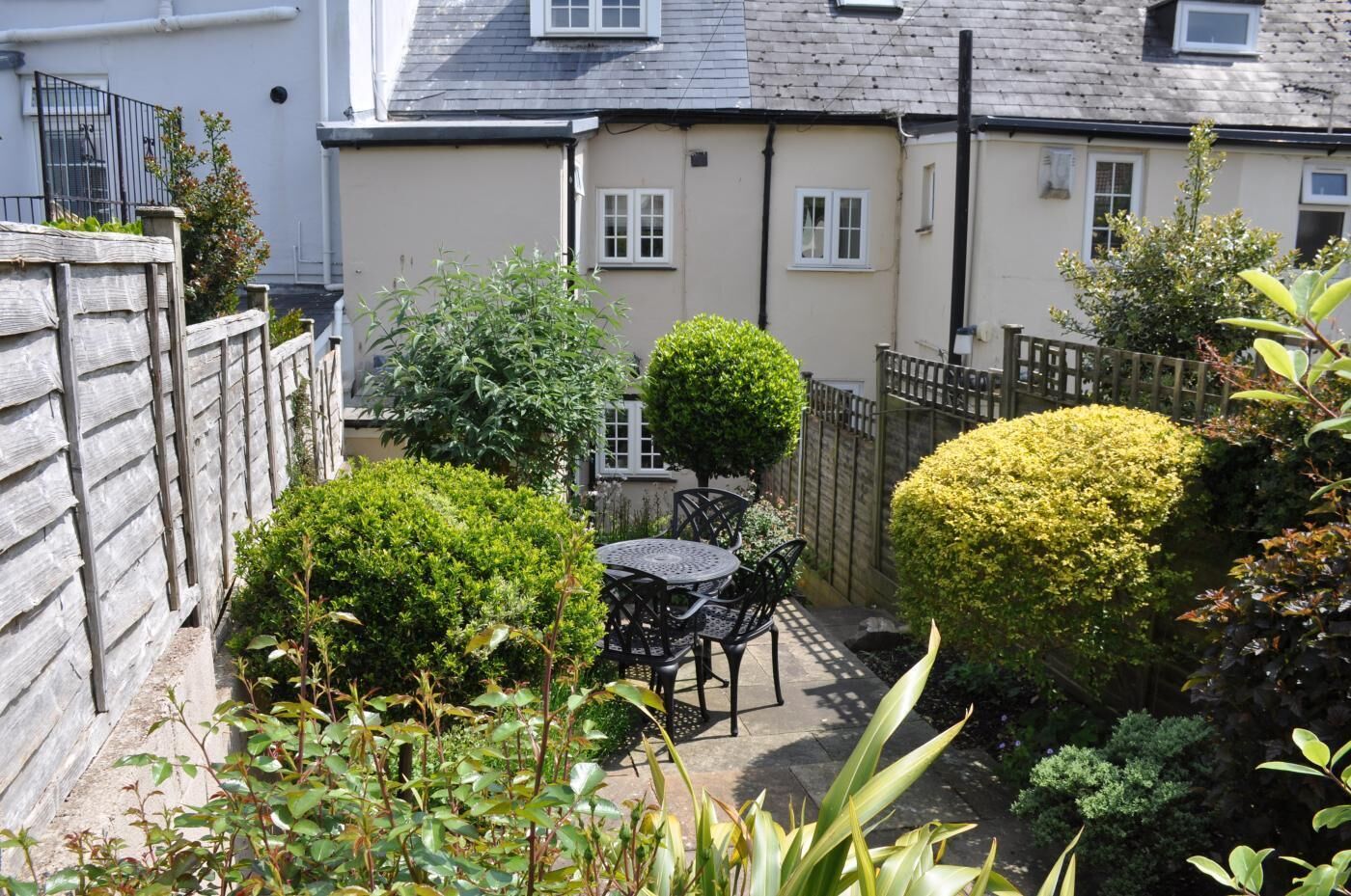 A small patio area with a round table and chairs, surrounded by various shrubs and plants.