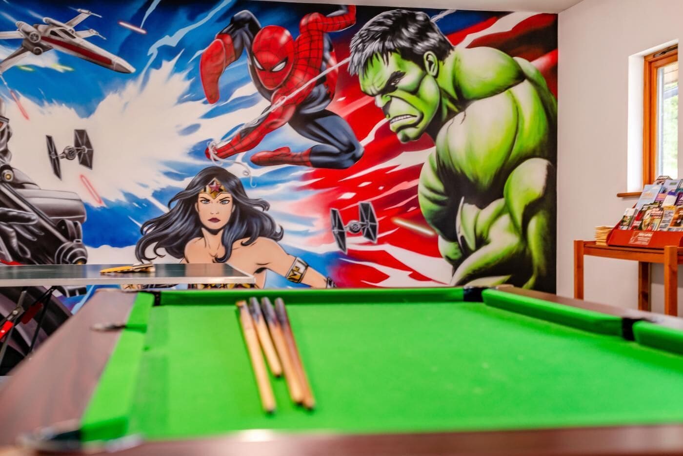 A pool table with a mural featuring superheroes and a table tennis paddle.