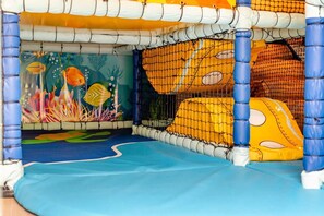 Ein Spielbereich für Kinder mit Netzumrandung, bunten Wandmalereien und weichen Spielelementen.