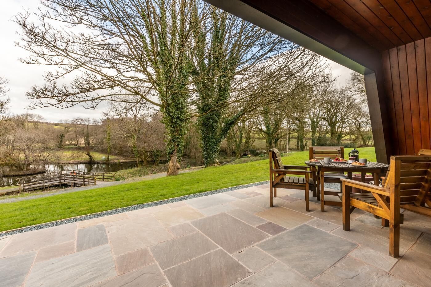 An outdoor patio with stone flooring, wooden furniture, and a view of a lake and trees.