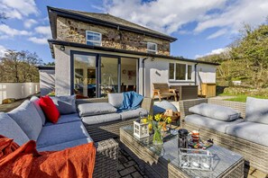 Une maison moderne avec une façade en pierre, de grandes portes vitrées donnant sur un patio aménagé avec des meubles d’extérieur et une table en verre.