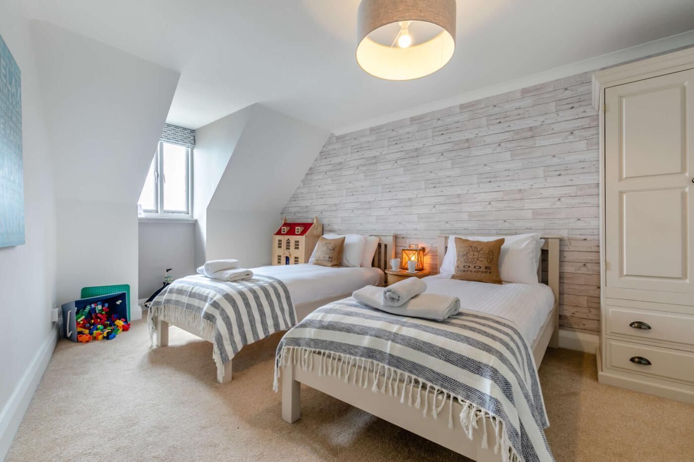 A bedroom with two beds, a wooden wall, a window, and a small play area with toys.