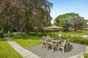 Een terras met tafel en stoelen, omgeven door een tuin met bomen en een grindpad.