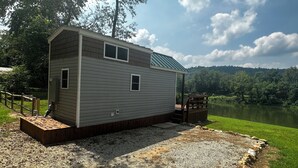 A small, single-story cabin with a metal roof, situated by a lake with a wooden deck and steps leading to a covered porch. - La Petite Maison, Tiny house Melbourne, AR in peaceful community of Mount Olive. (Melbourne)