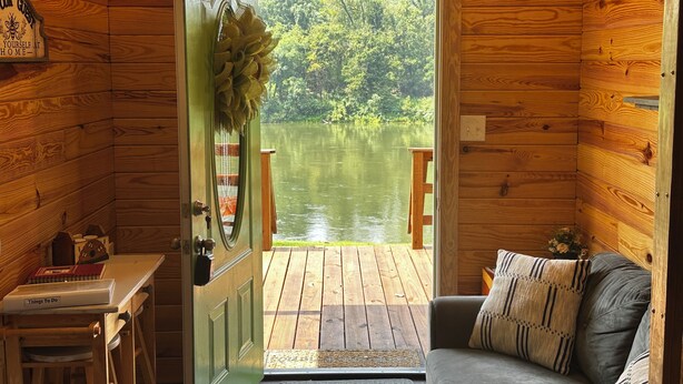 A cabin with a wooden interior, a green door leading to a dock over water, a small table with a book, and a couch with pillows. - La Petite Maison, Tiny house Melbourne, AR in peaceful community of Mount Olive. (Melbourne)