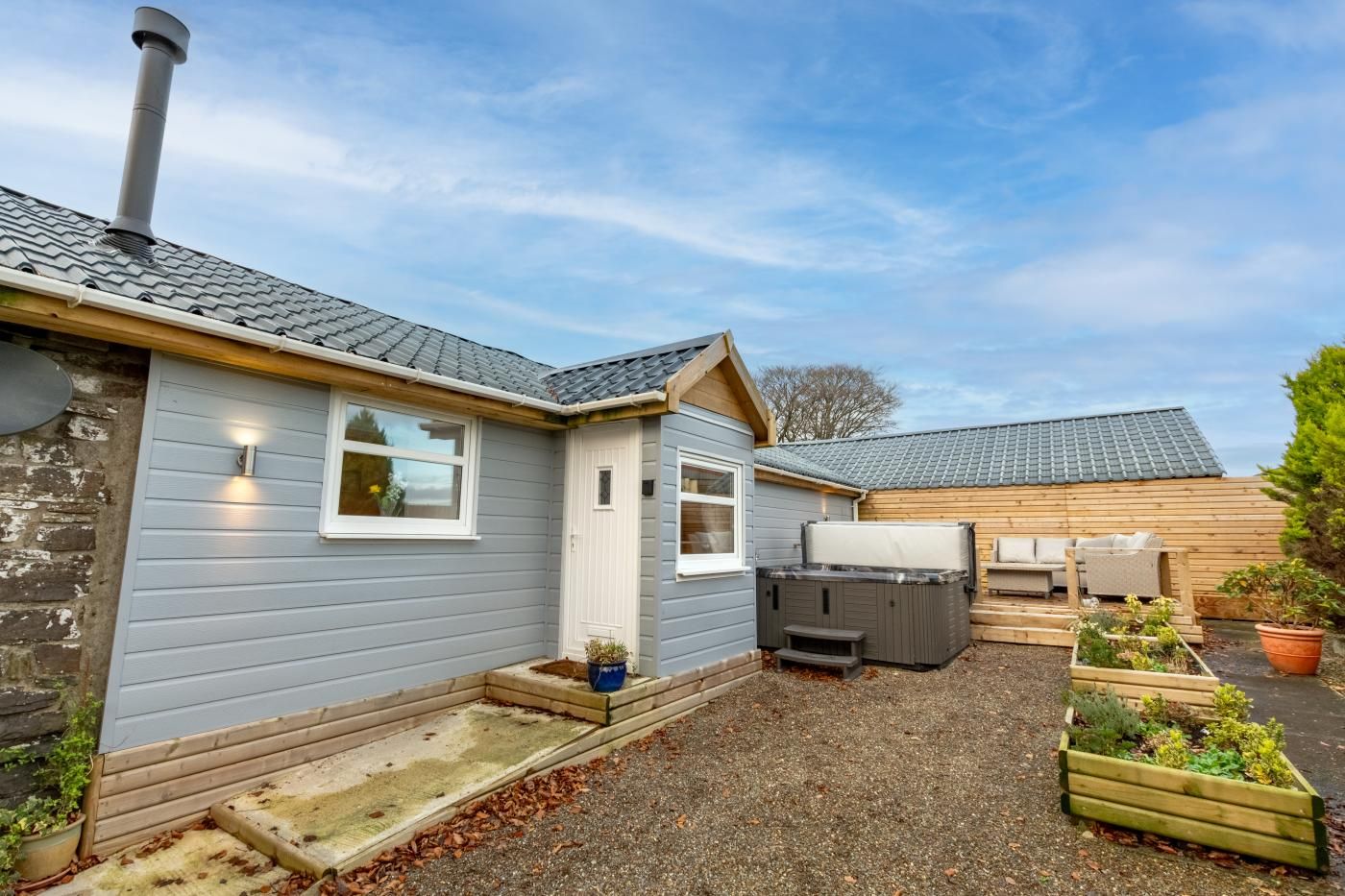A small, single-story house with a grey exterior, a chimney, a hot tub, and a wooden deck with potted plants.