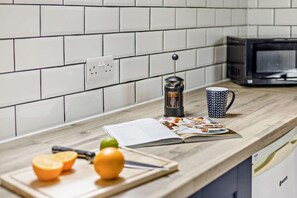 Un comptoir de cuisine avec une machine à café, une tasse à motifs, un magazine et des fruits sur une planche à découper.