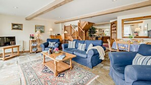 Un salon spacieux avec un grand canapé bleu, une table basse en bois et un tapis à motifs. Un escalier mène à un espace cuisine situé au fond de la pièce.