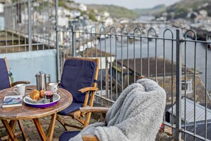 Une table avec une cafetière, des croissants et un livre, le tout placé devant un paysage de port avec des bateaux et une clôture en métal.