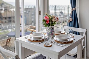Une table à manger dressée avec des assiettes, des couverts et un vase de fleurs, offrant une vue sur la ville à travers de grandes fenêtres.